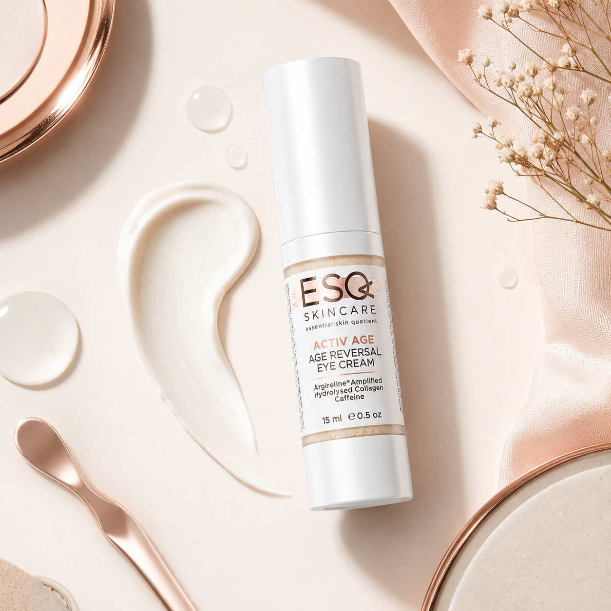 A bottle of ESQ Skincare Activ Age Age Reversal Eye Cream is displayed in a styled flat-lay arrangement with a white cream smear, rose gold-toned dishes, a spoon, and dried baby's breath on a soft beige background.