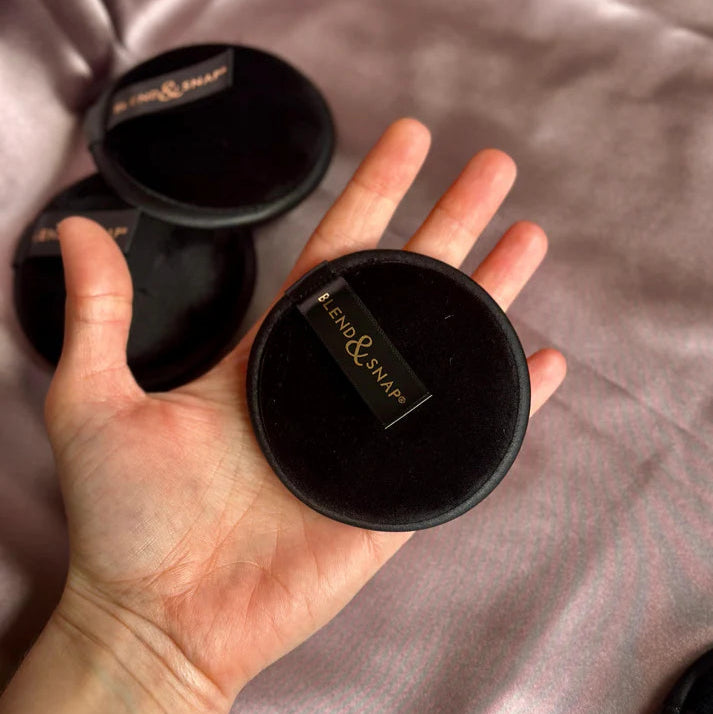 Hand holding a black compact mirror with 'Bentley & Sons' branding on a soft pink fabric background.