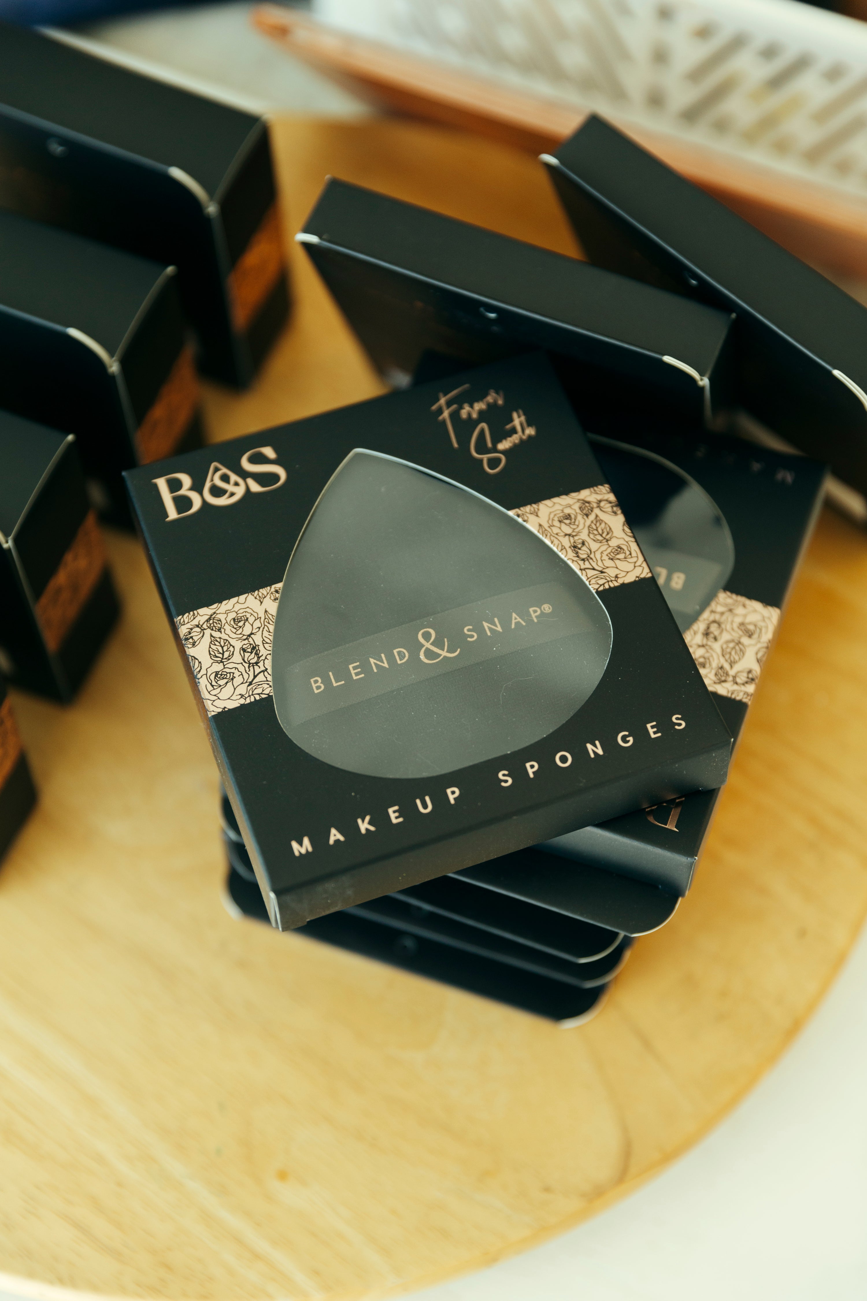 A stack of black B&S Blend & Snaps makeup sponges boxes arranged on a light wooden surface, with one box in sharp focus showing the product packaging design.