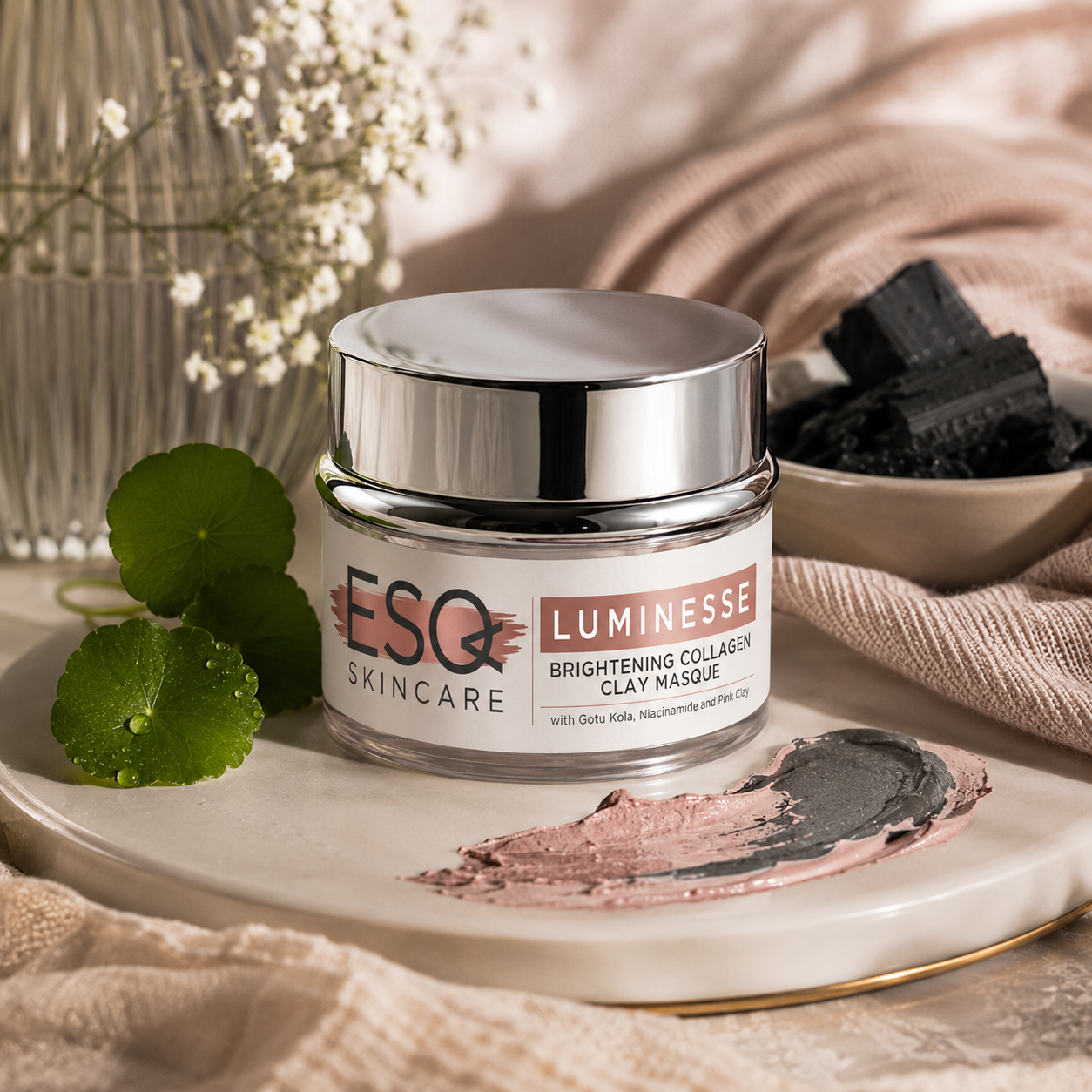 A jar of ESQ Skincare Luminesce Brightening Collagen Clay Masque sits on a ceramic tray with a smear of the dark clay mask. The scene is styled with green clemmint leaves, dried flowers, charcoal, and beige linen fabric.