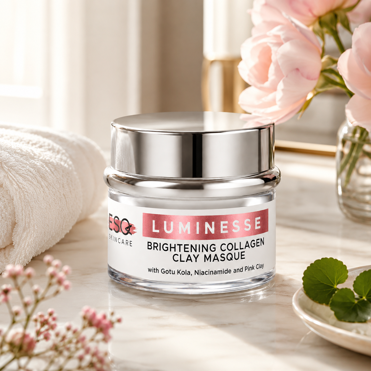 A jar of Luminesse Brightening Collagen Clay Masque skincare product on a marble surface, styled with a white towel, pink flowers, and green leaves.