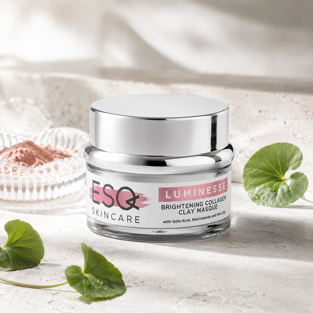 A jar of ESQ Skincare Luminesse Brightening Collagen Clay Masque, featuring a shiny silver lid and a white label with pink accents, styled on a textured light stone surface with green leaves and a decorative dish in the background.