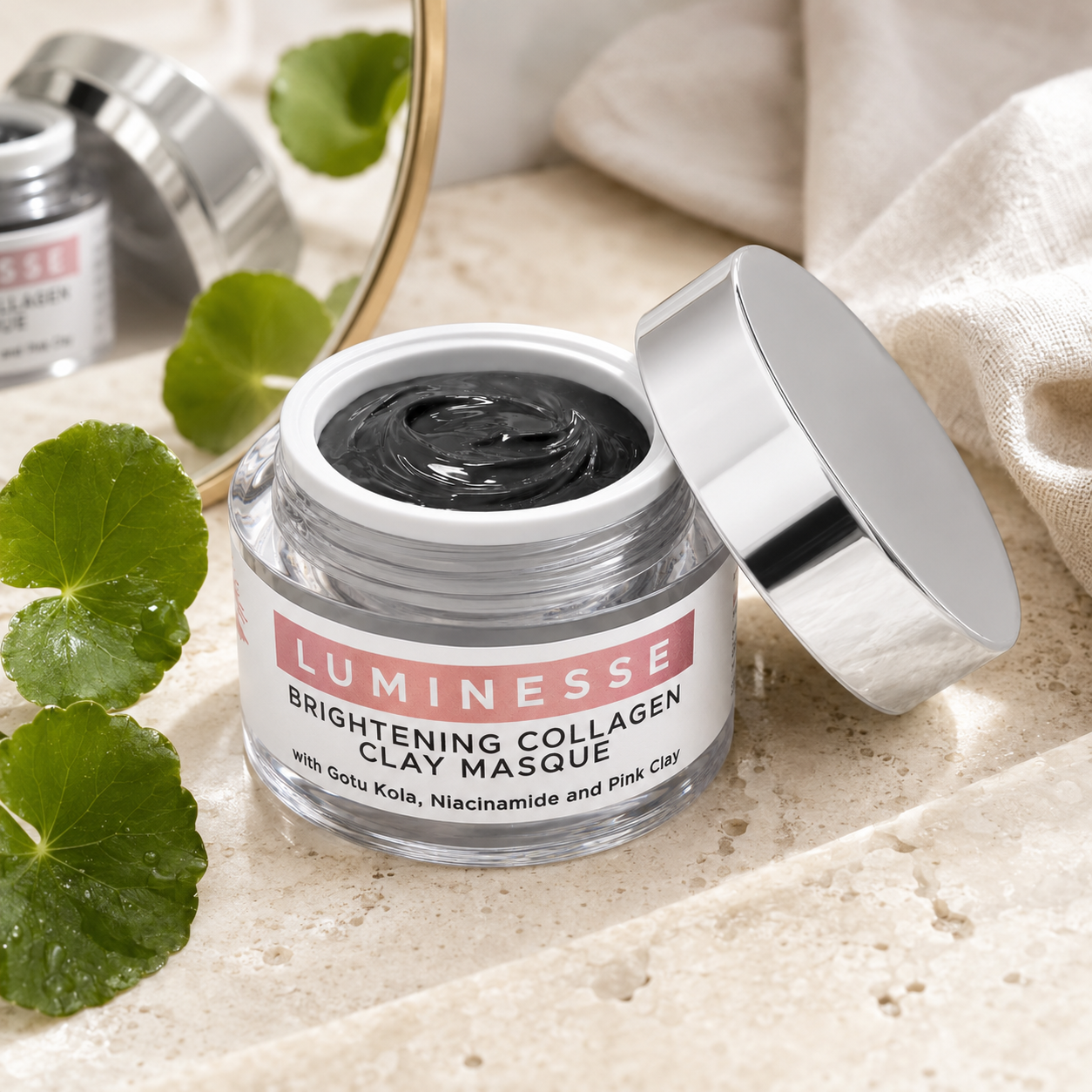An open jar of Luminesse Brightening Collagen Clay Masque showing the dark clay mask inside, with its silver lid resting beside it on a textured stone surface next to green leaves and a beige cloth.