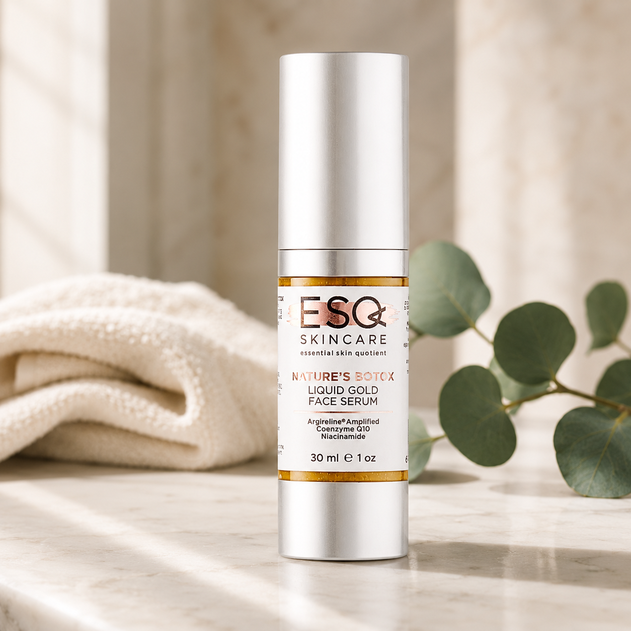 ESQ Skincare Nature's Botoxx Liquid Gold Face Serum in a silver cylindrical bottle with a white cap, set on a marble surface with a folded cream towel and green eucalyptus leaves in the background.