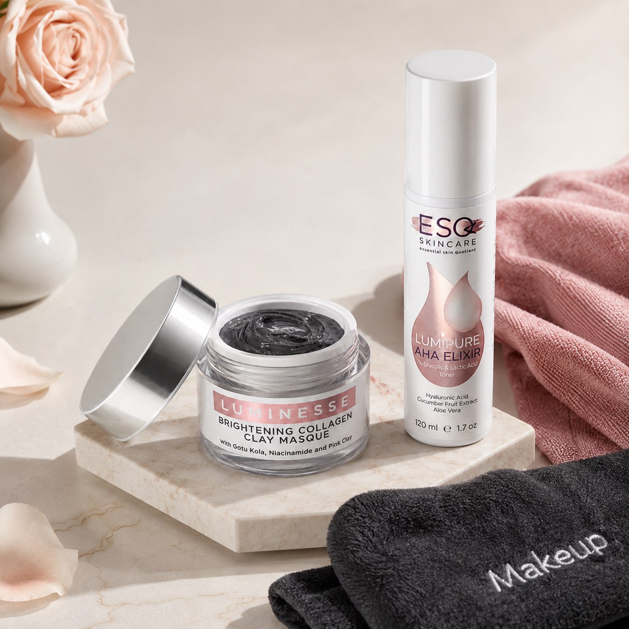 Two skincare products displayed on a marble tray: a jar of Lucinisse Brightening Collagen Clay Masque with its silver lid open, and a bottle of ESQ Skincare Lumi Pure AHA Energy. The scene is styled with a pink rose in a vase, a pink towel, and a black towel with the word 'Makeup' on it, set against a warm beige background.
